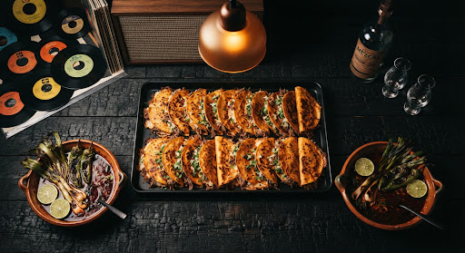Twenty-four crisp quesabirria tacos in a catering tray with two tubs of consommé, chiles toreados, and lime.