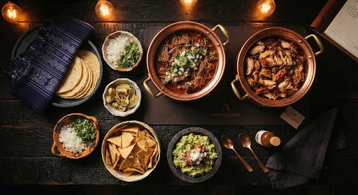 Overhead taco bar spread: birria and carnitas trays, tortillas, onion, cilantro, lime, totopos, and guacamole.