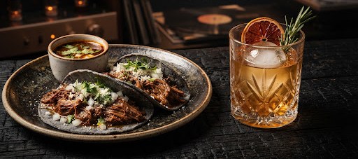 Birria tacos on blue corn tortillas beside a mezcal cocktail on the cantina table.